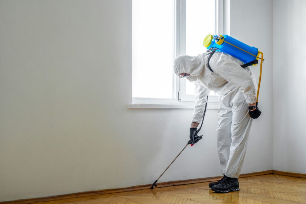 A pest control technician in protective gear spraying pesticide indoors with a backpack sprayer.